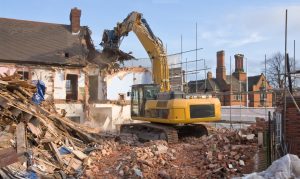"A large tracked excavator machine fitted with a demolition tool, ripping apart old houses and buildings in a town centre for re-development."